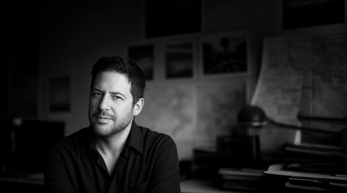 Black-and-white portrait of Robin Bucciarelli in a dark shirt, seated in his studio and looking into the camera, framed prints and a paper-covered desk behind him.